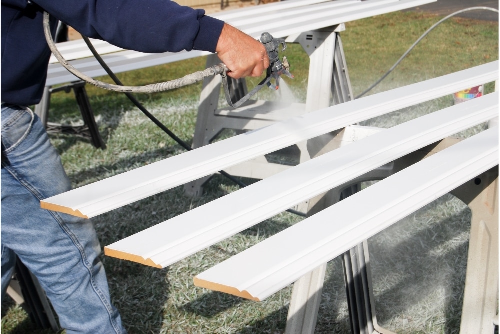 A person spray painting wood trim in white outdoors.