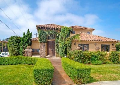 A two-story house with a red tile roof, surrounded by greenery and hedges.
