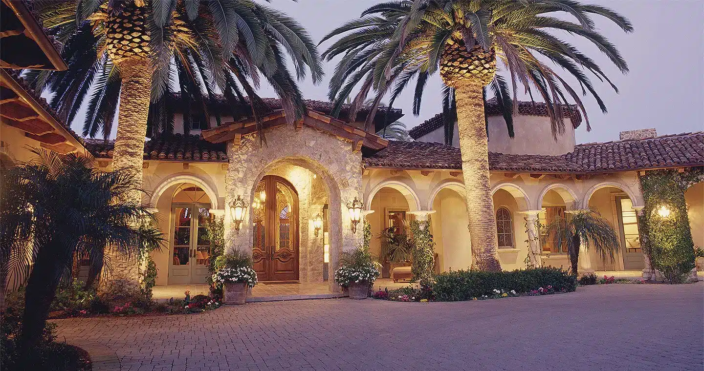 A landscaped entrance to a luxurious home with palm trees and stone architecture.