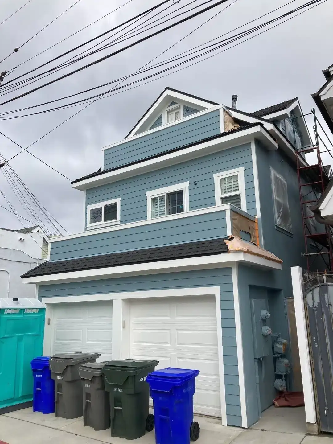 Two-Story Blue House with Garage Exterior of a two-story house with a blue facade and garage doors, featuring garbage bins in front.
